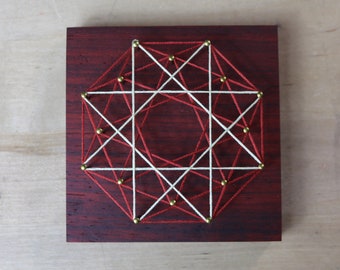 May include: A geometric design created with red and white string wrapped around gold tacks on a dark wood background.