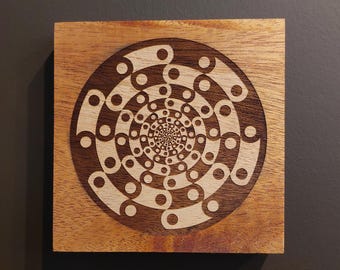 May include: Square wooden art piece with a spiral design. The artwork features a light-colored, intricate pattern etched into a darker wood background. The design is centered on the square, creating a symmetrical and visually appealing piece.