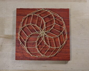 May include: A geometric design created with string wrapped around nails hammered into a wooden board. The string is a light brown color and the nails are silver. The board is a dark red color.