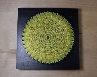 May include: A yellow string art piece in the shape of a sun on a black wooden background.