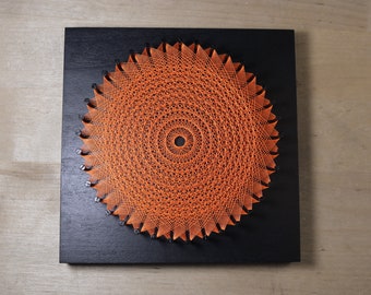 May include: Orange string art on a black wooden square. The string is woven in a circular pattern, creating a sunburst design. The artwork is mounted on a black wooden base, with small nails securing the string. A unique piece of wall decor.