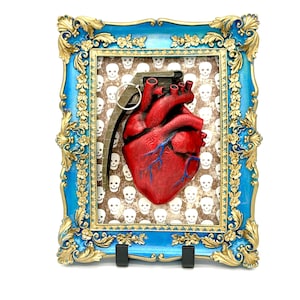 May include: A framed artwork featuring a red anatomical heart and a grenade design. The heart is detailed with blue veins, set against a backdrop of white skulls. The frame is ornate, with a blue and gold color scheme.