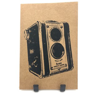 May include: A black and white image of a vintage Kodak Duplex Camera with the text "Kodak Lens" and "Kodak Duplex Camera" printed on the front. The camera is printed on a brown paper background.