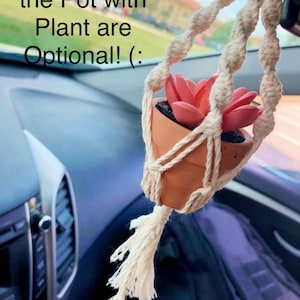 May include: A white macrame hanging with a small terracotta pot and a pink artificial succulent plant. The hanging is attached to a car's rearview mirror. The text "**Charms and the Pot with Plant are Optional! :" is visible in the image.