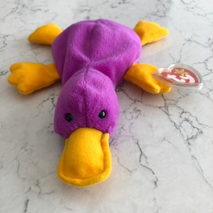 Può includere: Giocattolo di peluche platypus viola e giallo con un'etichetta Ty rossa e bianca.