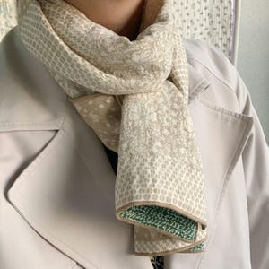 May include: A beige scarf with a floral and dot pattern is draped over a light beige coat. The scarf has a green border on one side. The coat has a button closure.