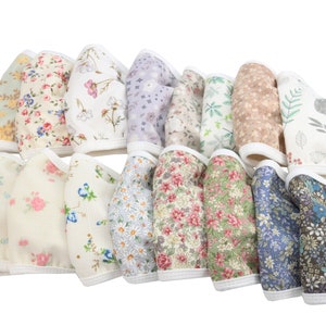 Op de afbeelding: Een collectie van zestien stoffen gezichtsmaskers gerangschikt in twee rijen, elk met een unieke bloemen- of botanische print in diverse kleuren waaronder roze, blauw, groen en bruin, afgewerkt met een witte rand en bijpassende witte oorlussen.