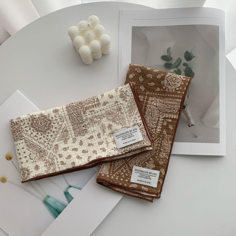 May include: Two rectangular fabric cloths featuring a brown and white paisley pattern. One cloth is primarily cream with brown print, while the other is brown with white print. Each has a white label reading HANDMADE BY AVO.