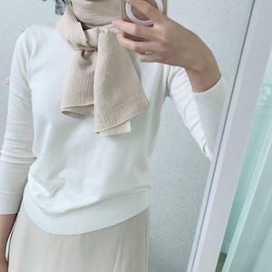 Peut inclure: Un pull blanc &agrave; manches longues associ&eacute; &agrave; une &eacute;charpe beige textur&eacute;e nou&eacute;e autour du cou. L'&eacute;charpe pr&eacute;sente un motif quadrill&eacute; visible et est port&eacute;e sur le pull, qui poss&egrave;de une texture en maille lisse et des poignets c&ocirc;tel&eacute;s.