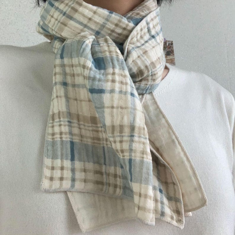 Peut inclure: Une &eacute;charpe l&eacute;g&egrave;re pr&eacute;sentant un motif &agrave; carreaux dans des tons cr&egrave;me, beige et bleu doux. Le tissu poss&egrave;de une texture froiss&eacute;e et douce avec des coutures visibles sur les bords, nou&eacute;e simplement autour du cou sur un haut blanc uni.