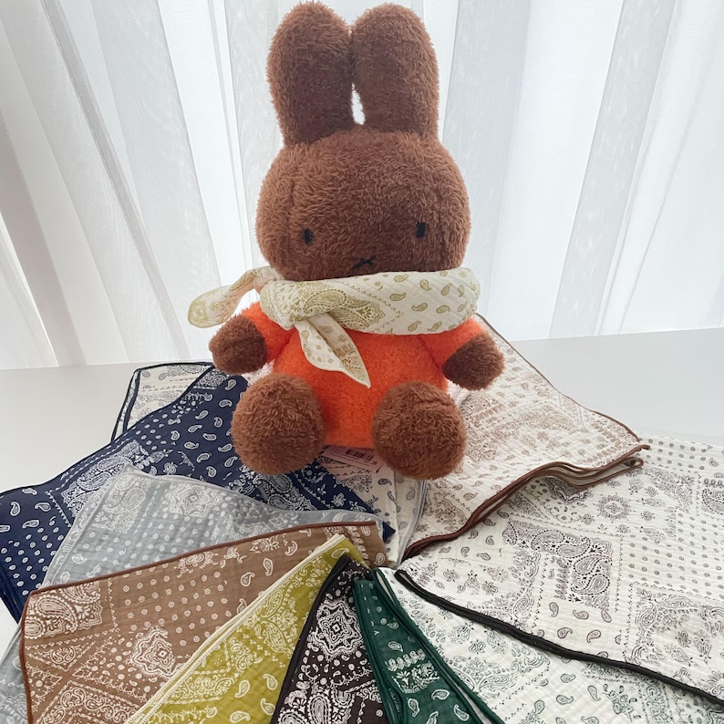 Puede incluir: Un juguete de peluche de conejo marr&oacute;n con orejas largas, que lleva una camisa naranja y un pa&ntilde;uelo color crema con estampado de cachemira alrededor del cuello, colocado sobre una colecci&oacute;n de pa&ntilde;uelos de tela cuadrados con varios patrones.