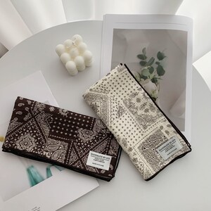 May include: Two folded rectangular fabric cloths featuring a dark brown and a cream-colored paisley bandana pattern. Each cloth has a white rectangular fabric label stitched to the corner that reads HANDMADE BY AVO SIMPLY MADE WORTH KEEPING MADE WITH CARE.