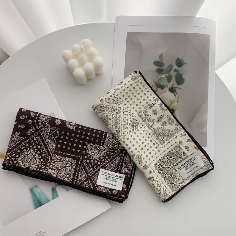 May include: Two folded rectangular fabric cloths featuring a dark brown and a cream-colored paisley bandana pattern. Each cloth has a white rectangular fabric label stitched to the corner that reads HANDMADE BY AVO SIMPLY MADE WORTH KEEPING MADE WITH CARE.
