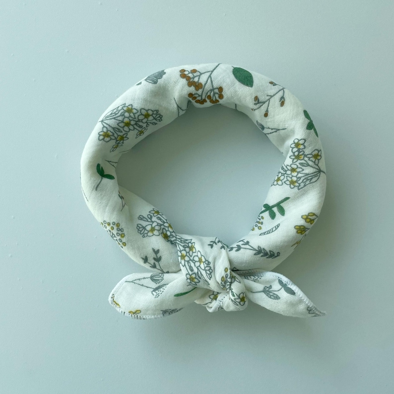 May include: A white fabric bandana tied in a circular knot, featuring a repeating botanical print of small green leaves, thin grey stems, and clusters of tiny yellow flowers scattered across the soft, textured material of the accessory.