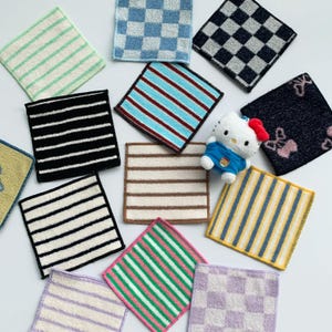 May include: A collection of square fabric cloths featuring various patterns including stripes and checkers in colors like blue, black, yellow, green, and purple, alongside a small white cat figurine wearing a blue shirt and a red bow.