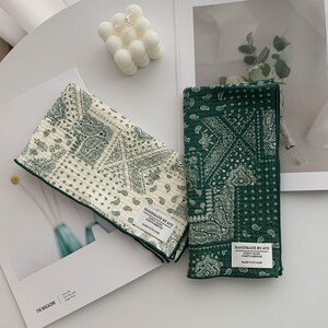 May include: Two rectangular fabric cloths featuring a green and white paisley pattern. One cloth is primarily white with green print, while the other is green with white print. Each has a white label reading HANDMADE BY AVO.