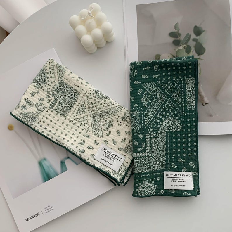 May include: Two rectangular fabric cloths featuring a green and white paisley pattern. One cloth is primarily white with green print, while the other is green with white print. Each has a white label reading HANDMADE BY AVO.