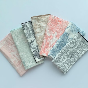 Pode incluir: Uma coleção de seis panos dobrados, com padrões e cores variadas, incluindo rosa, verde, bege, coral, azul e cinza. Cada pano apresenta um design detalhado e repetitivo. Os panos estão dispostos em forma de leque.