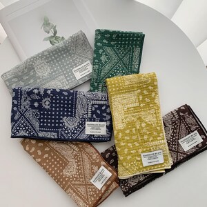 May include: A collection of six rectangular fabric cloths featuring a white paisley and geometric bandana pattern. The cloths come in gray, dark green, navy blue, mustard yellow, tan, and dark brown, each with a white label reading HANDMADE BY AVO.