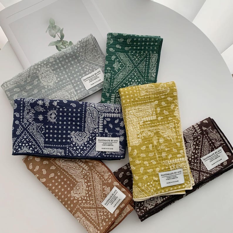 May include: A collection of six rectangular fabric cloths featuring a white paisley and geometric bandana pattern. The cloths come in gray, dark green, navy blue, mustard yellow, tan, and dark brown, each with a white label reading HANDMADE BY AVO.