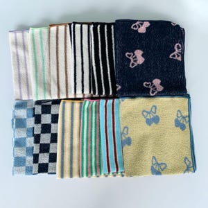 May include: A collection of folded washcloths in various patterns and colours. Designs include stripes, checks, and floral motifs. The colour palette includes blue, black, white, yellow, and pink. The washcloths are neatly arranged in a grid-like formation.