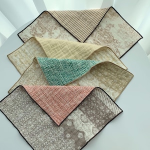 May include: A collection of folded, patterned fabric squares in various colors. The fabrics feature designs in beige, green, and pink, with black trim. The patterns include stripes, floral motifs, and abstract designs. The squares are arranged on a white surface.