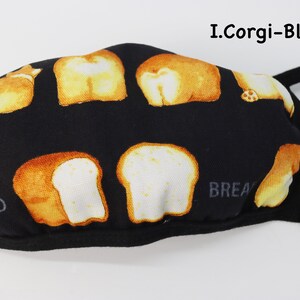 Op de afbeelding: Een zwart mondkapje met een herhalend patroon van goudbruine corgi-achterwerken en sneetjes witbrood. De tekst I.Corgi-Black staat in de rechterbovenhoek en het woord BREAD is in het grijs zichtbaar aan de linker- en rechteronderkant.