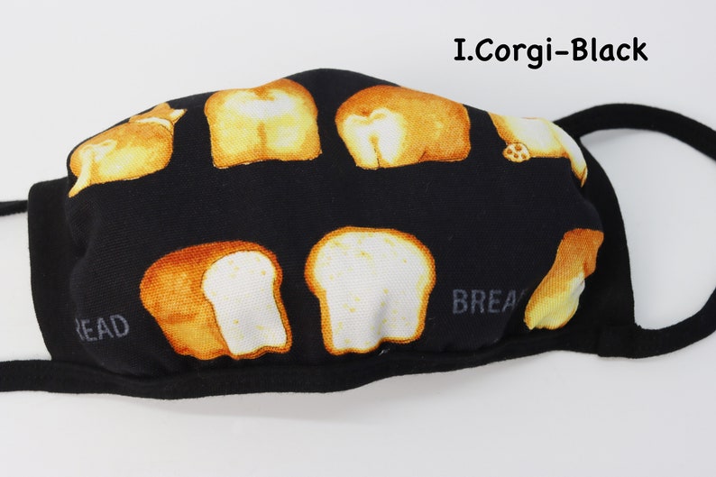 Op de afbeelding: Een zwart mondkapje met een herhalend patroon van goudbruine corgi-achterwerken en sneetjes witbrood. De tekst I.Corgi-Black staat in de rechterbovenhoek en het woord BREAD is in het grijs zichtbaar aan de linker- en rechteronderkant.