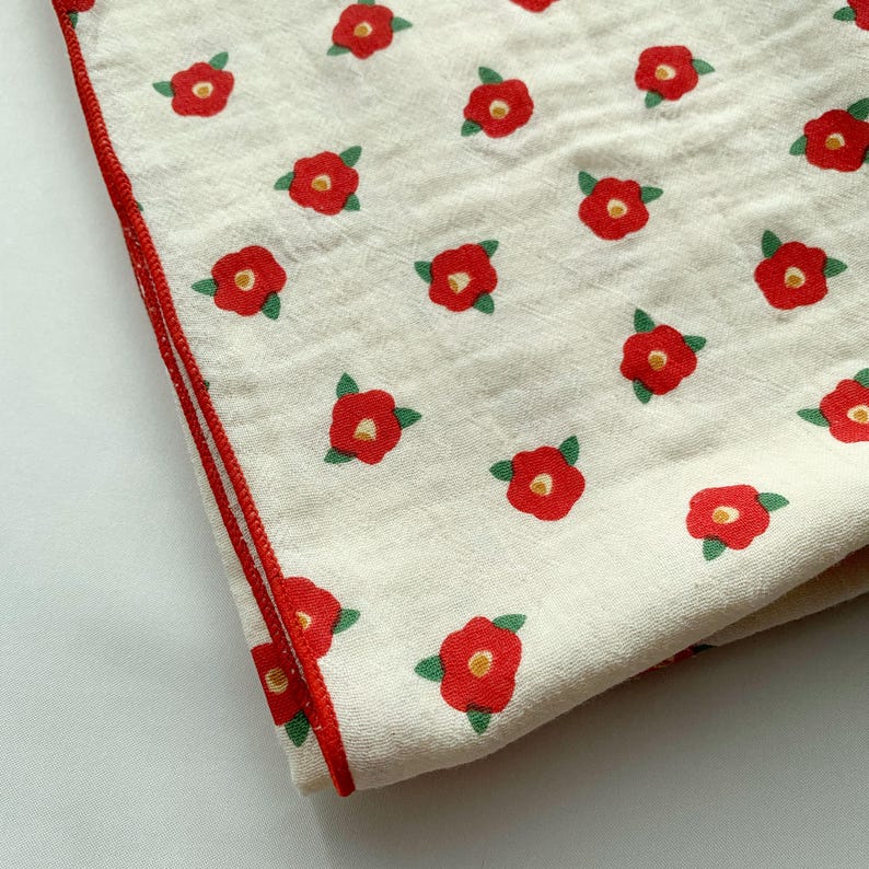 May include: A cream-colored fabric textile featuring a repeating pattern of small red flowers with yellow centers and green leaves. The fabric has a textured, layered appearance and is finished with a thin, vibrant red stitched border edge.