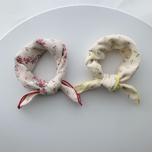 May include: Two square fabric scarves tied into knots. The left scarf is off-white with a dense pattern of small red roses and green leaves, finished with a red stitched edge. The right scarf is off-white with tiny yellow and green floral sprigs.