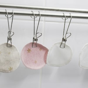 Puede incluir: Siete almohadillas redondas de algodón para desmaquillar colgadas de una cuerda de ropa plateada con pinzas. Las almohadillas son blancas, beige con diseños de copos de nieve dorados y rosas con diseños de copos de nieve dorados.