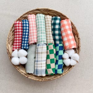 May include: A woven basket holds a collection of rolled, checkered fabric in various colors, including red, blue, pink, green, and orange. Fluffy cotton buds are placed near the fabric rolls. The basket is made of natural fibers.