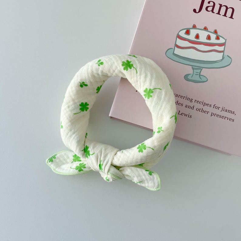 May include: A cream-colored fabric neckerchief featuring a repeating pattern of small green four-leaf clovers. The accessory is made of a textured, crinkled material and is tied into a circular loop with two short, pointed ends at the bottom.