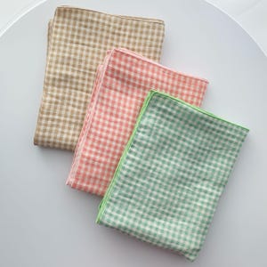 Könnte beinhalten: Drei gefaltete Stoffquadrate mit Vichy-Muster. Die Farben sind Beige, Koralle und Mintgrün. Das grüne Quadrat hat einen grünen Rand. Die Quadrate sind auf einer weißen Oberfläche angeordnet.