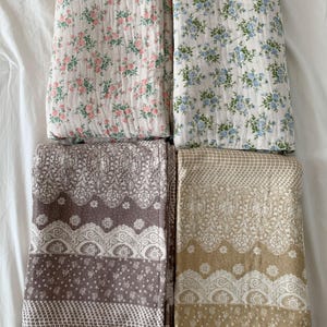 May include: Four folded rectangular blankets arranged in a grid. The top left features pink floral patterns on white, the top right has blue floral patterns on white, the bottom left is brown with white lace-like motifs, and the bottom right is tan.