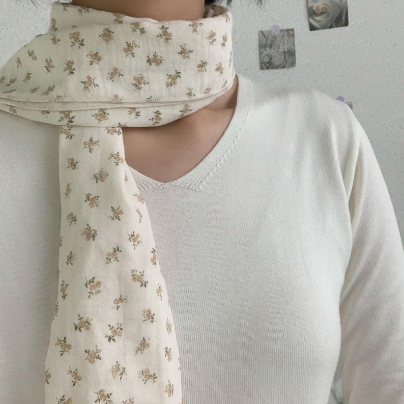 May include: A long, rectangular scarf made of off-white fabric featuring a repeating pattern of small, light brown floral sprigs. The scarf is draped around the neck, paired with a plain, long-sleeved, off-white V-neck sweater with a ribbed texture.