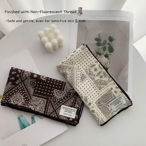 May include: Two folded square cloths featuring a dark brown and a cream paisley bandana pattern. Each cloth has a white rectangular label reading "HANDMADE BY AVO". Text on the image states: "Finished with Non-Fluorescent Thread -Safe and gentle, even for sensitive skin & kids".