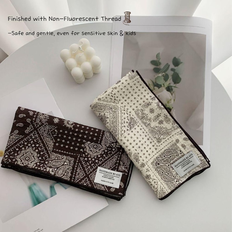 May include: Two folded square cloths featuring a dark brown and a cream paisley bandana pattern. Each cloth has a white rectangular label reading "HANDMADE BY AVO". Text on the image states: "Finished with Non-Fluorescent Thread -Safe and gentle, even for sensitive skin & kids".
