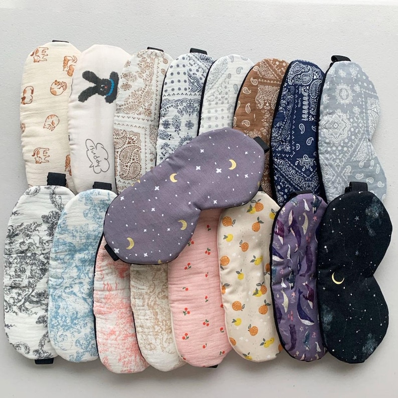 Op de afbeelding: Een verzameling van 16 slaapmaskers met verschillende patronen en kleuren. De maskers zijn gemaakt van zacht materiaal en hebben een zwart elastiek. Sommige van de patronen omvatten sterren, manen, citroenen, kersen en paisley.
