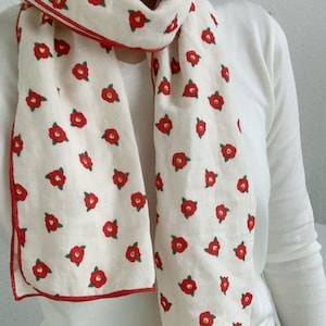 May include: A cream-colored scarf featuring a repeating pattern of small red flowers with green leaves. The scarf is finished with a thin red stitched border along the edges, providing a distinct contrast against the light fabric background.