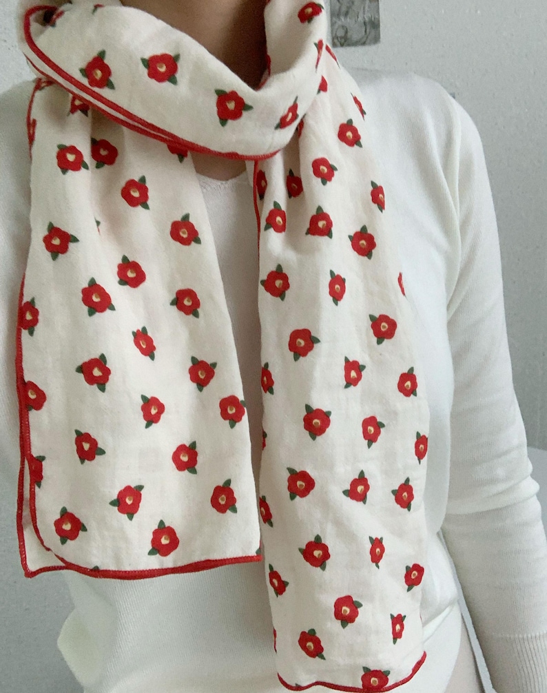 May include: A cream-colored scarf featuring a repeating pattern of small red flowers with green leaves. The scarf is finished with a thin red stitched border along the edges, providing a distinct contrast against the light fabric background.