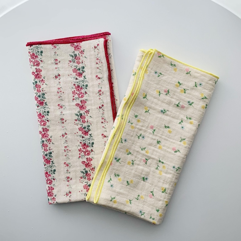May include: Two rectangular fabric cloths with a cream-colored base. One cloth features vertical stripes of pink and red floral patterns with a red stitched edge. The other cloth displays a scattered pattern of tiny yellow and blue flowers with a yellow stitched edge.