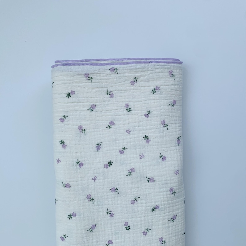 May include: A folded piece of white fabric with a textured, quilted appearance. The textile features a repeating pattern of small, light purple flowers with tiny green stems and leaves. The top edge is finished with a light purple fabric trim.