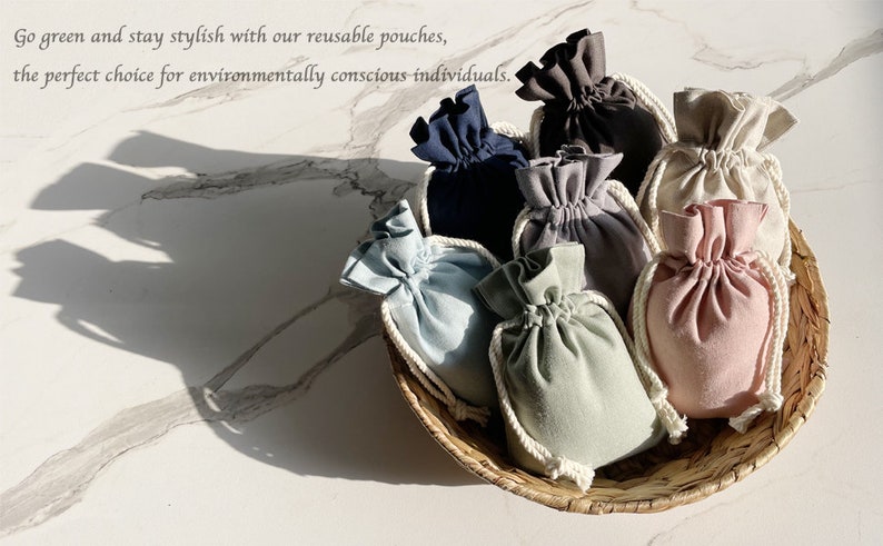 K&ouml;nnte beinhalten: Eine Sammlung wiederverwendbarer Stoffbeutel in verschiedenen Farben, darunter hellblau, grau, beige und rosa, die in einem geflochtenen Korb angeordnet sind. Die Beutel sind aus weichem, nat&uuml;rlichem Stoff gefertigt und haben einen Kordelverschluss.