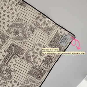 May include: A quilted fabric piece featuring a black and white paisley bandana pattern with a black border. A small white rectangular label is sewn on the corner with the text "HANDMADE BY AYO" and "MADE WITH CARE". A pink arrow points to the label.