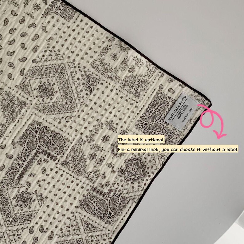 May include: A quilted fabric piece featuring a black and white paisley bandana pattern with a black border. A small white rectangular label is sewn on the corner with the text "HANDMADE BY AYO" and "MADE WITH CARE". A pink arrow points to the label.
