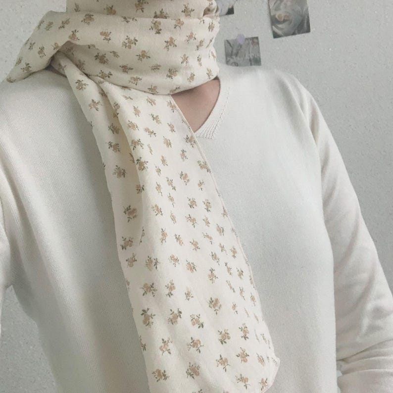 Peut inclure: Une &eacute;charpe de couleur cr&egrave;me orn&eacute;e d'un motif r&eacute;p&eacute;titif de petites fleurs marron clair, port&eacute;e sur un pull &agrave; manches longues et col en V de couleur cr&egrave;me. L'&eacute;charpe est drap&eacute;e autour du cou, avec une extr&eacute;mit&eacute; tombant sur le devant du torse.