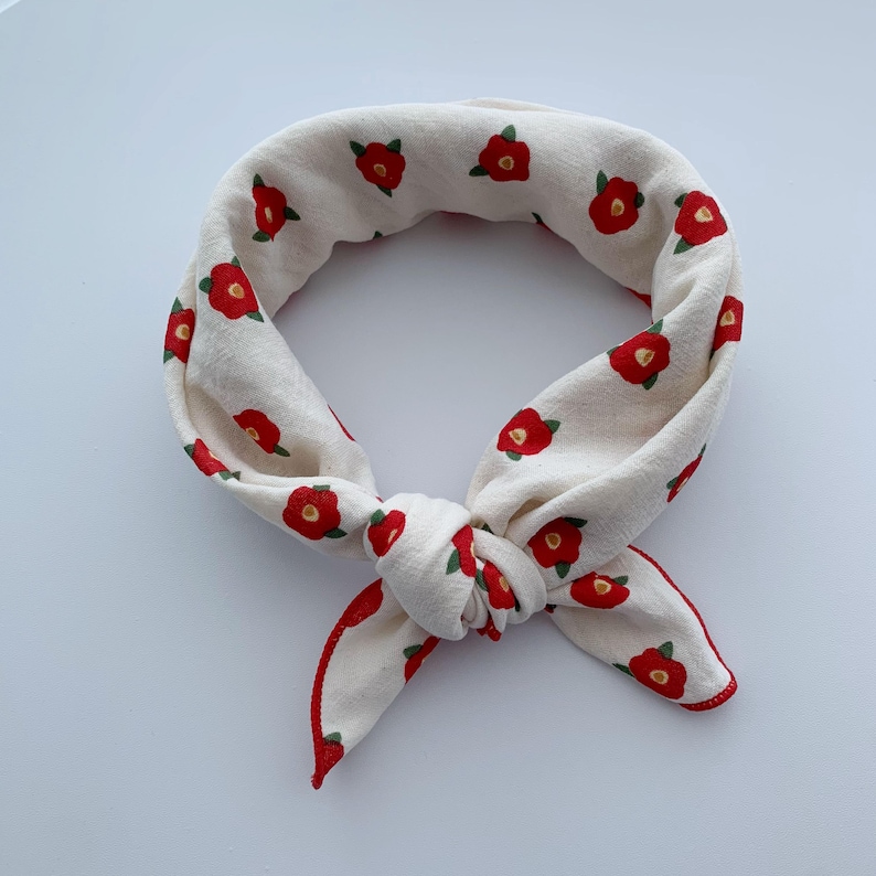 May include: A cream-colored fabric neckerchief or bandana featuring a repeating pattern of small red camellia flowers with green leaves. The accessory is tied into a circular loop with a knot at the bottom, showcasing a red stitched hem edge.
