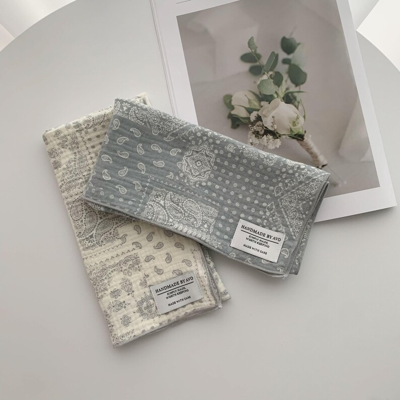 May include: Two rectangular cotton cloths featuring a grey and white paisley pattern. Each cloth has a quilted texture and a white fabric label stitched to the corner that reads HANDMADE BY AVO, SIMPLY MADE, WORTH KEEPING, MADE WITH CARE.