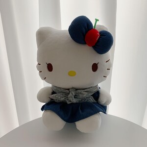 May include: A white plush character toy featuring a large dark blue denim bow with a red cherry accent on its head. The character wears a dark blue denim dress with a grey patterned neck scarf. It has a yellow nose, black eyes, and whiskers.